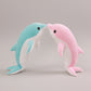 Blue and pink dolphin plush toys standing nose to nose on a light neutral background