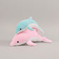 Blue dolphin plush toy resting on a pink dolphin plush toy on a light neutral background