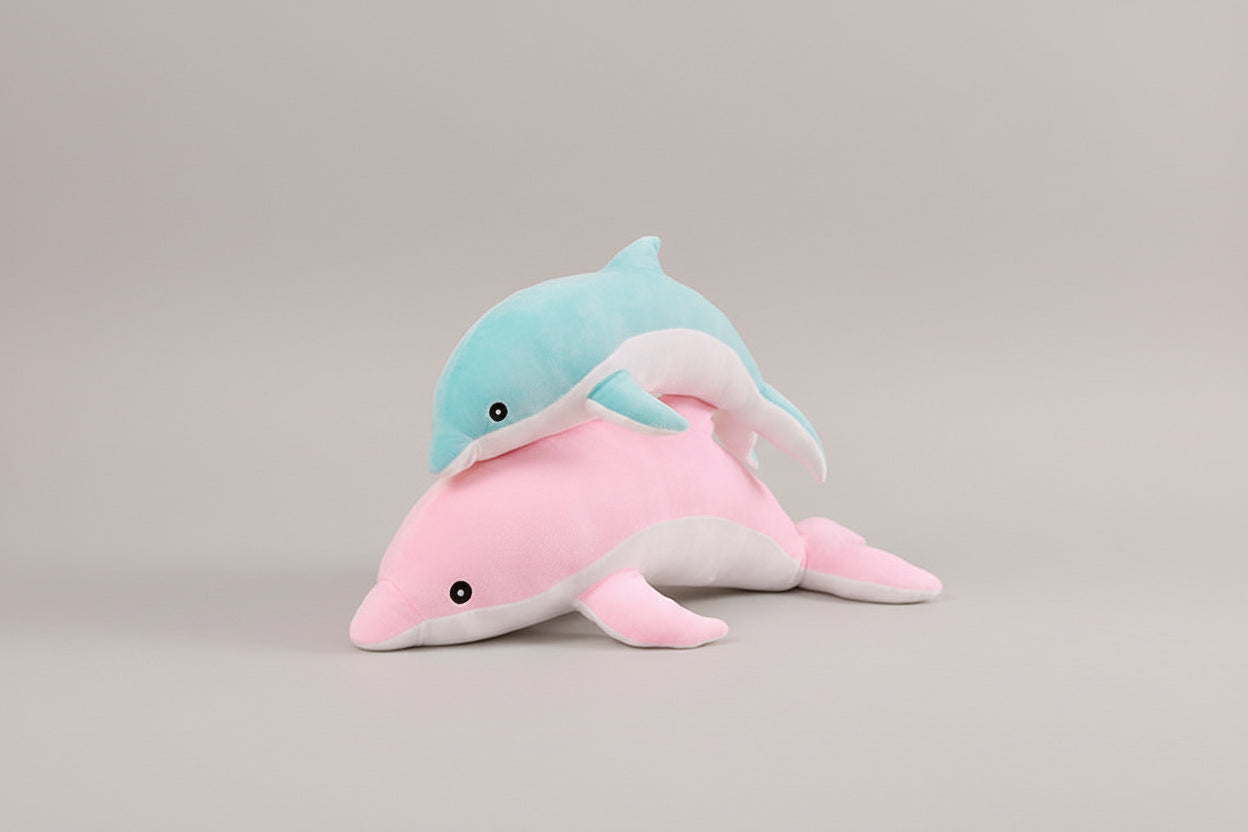 Blue dolphin plush toy resting on a pink dolphin plush toy on a light neutral background