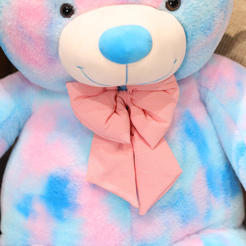 Close up of a pastel teddy bear plush toy featuring pink and blue tie dye colours with a decorative bow