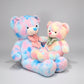 Two pastel teddy bear plush toys in pink blue and rainbow colours sitting together on a neutral background