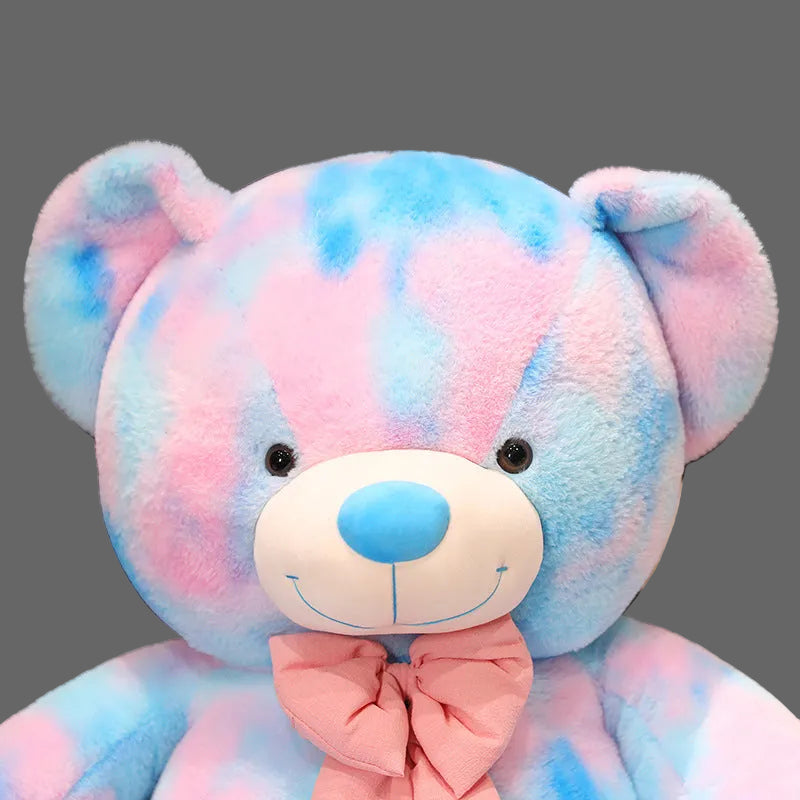 Pastel teddy bear plush toy with pink and blue tie dye pattern and decorative bow on a grey background