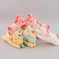 Pastel unicorn plush toy collection in pink, white, yellow and green colours on a light grey background.