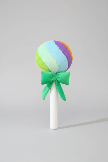 Rainbow lollipop plush toy with green ribbon bow on a light grey background.