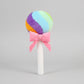 Rainbow lollipop plush toy with pink ribbon bow on a light grey background.