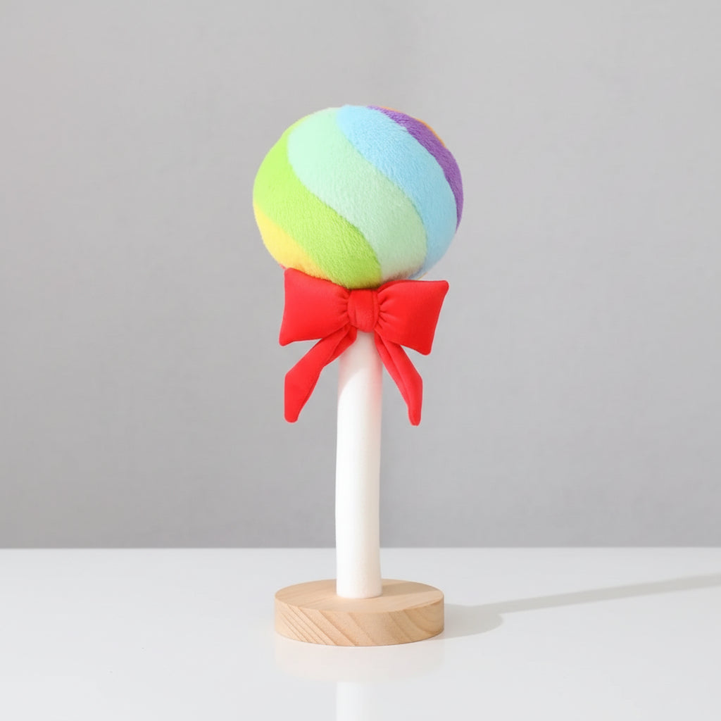 Rainbow lollipop plush toy with red ribbon bow displayed on a wooden stand against a light grey background.