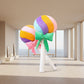 Rainbow lollipop plush toys with colourful bows standing in a modern living room with floor-to-ceiling windows.