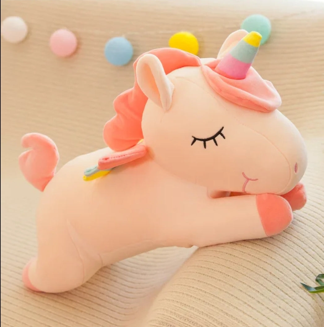 Sleeping unicorn plush toy with pastel rainbow horn, pink mane and rainbow wings resting on a soft cushion.