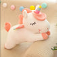 White sleeping unicorn plush toy with pastel rainbow horn, pink mane and rainbow wings resting on a soft knitted cushion.