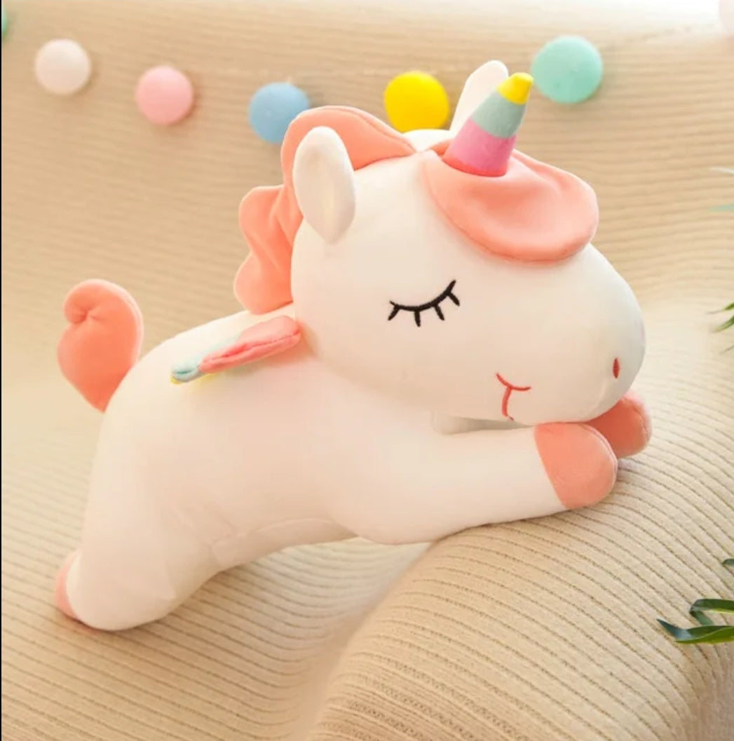 White sleeping unicorn plush toy with pastel rainbow horn, pink mane and rainbow wings resting on a soft knitted cushion.