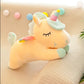 Yellow sleeping unicorn plush toy with pastel rainbow horn, rainbow wings and mint accents resting on a soft knitted cushion.